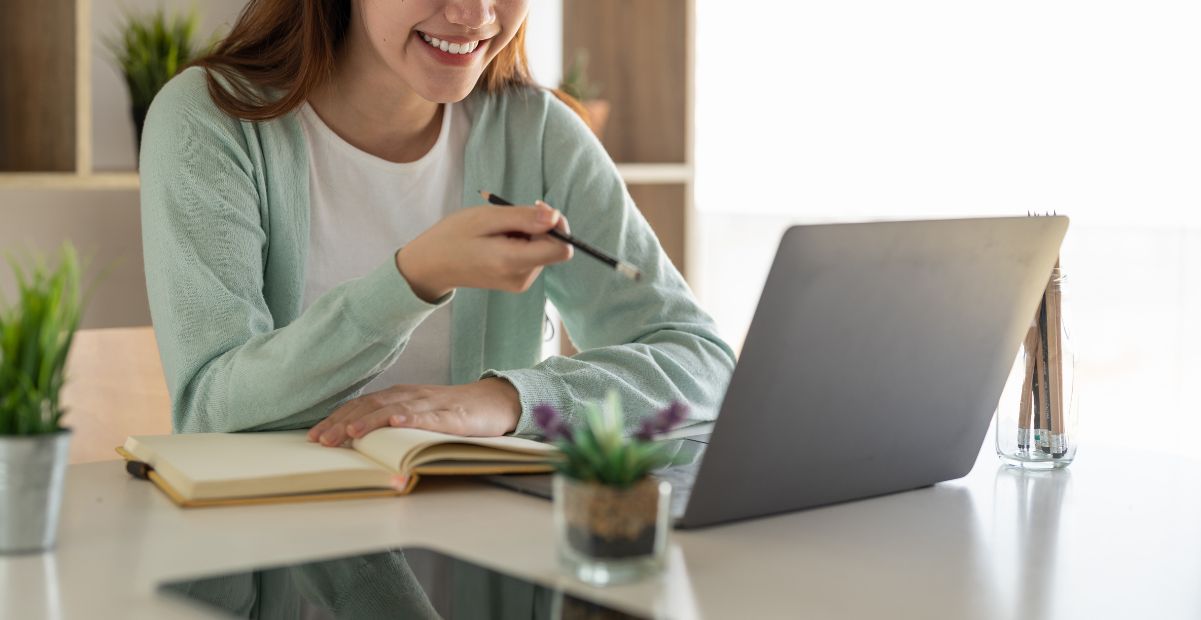 Mulher sorrindo trabalhando em um ambiente acolhedor com laptop e caderno, representando a integração de saberes e práticas no empreendedorismo feminino.