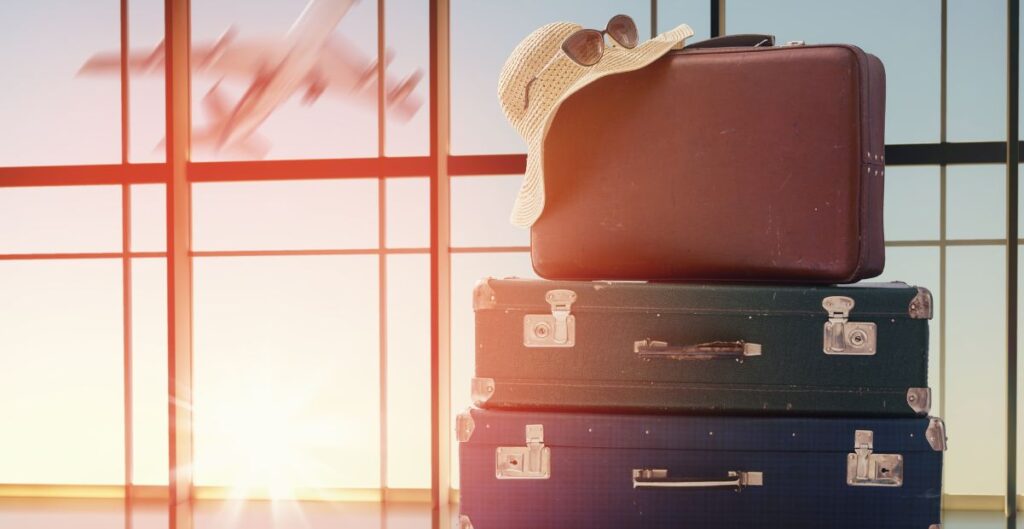 Malas de viagem empilhadas com um chapéu e óculos de sol em um aeroporto, com um avião ao fundo.