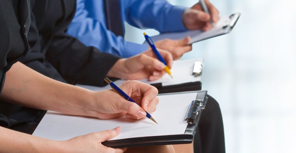 Pessoas escrevendo em pranchetas durante um treinamento profissional, simbolizando postura e preparo para entrevistas de emprego.