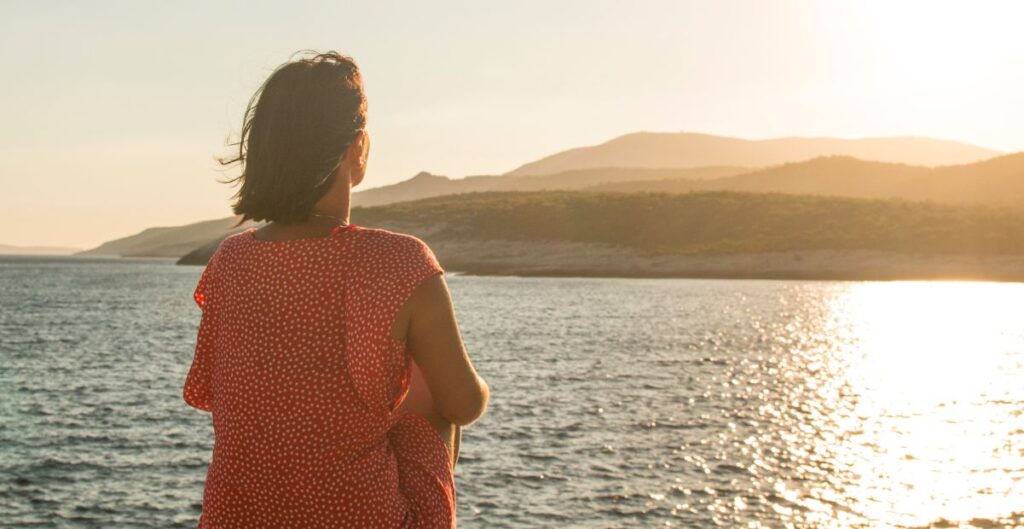 Mulher olhando para o horizonte ao pôr do sol, refletindo à beira do mar, representando um momento de pausa e introspecção.