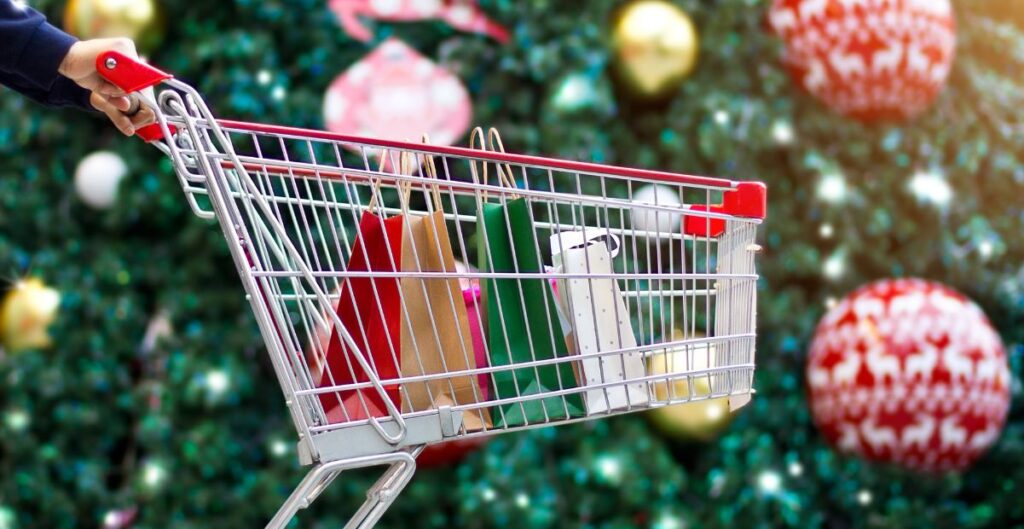 Carrinho de compras com sacolas coloridas em frente a uma decoração natalina.