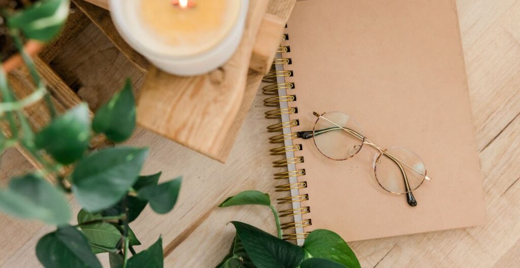 Caderno com óculos de leitura sobre uma mesa de madeira, cercado por plantas e uma vela acesa, simbolizando organização e bem-estar no contexto da economia do cuidado.