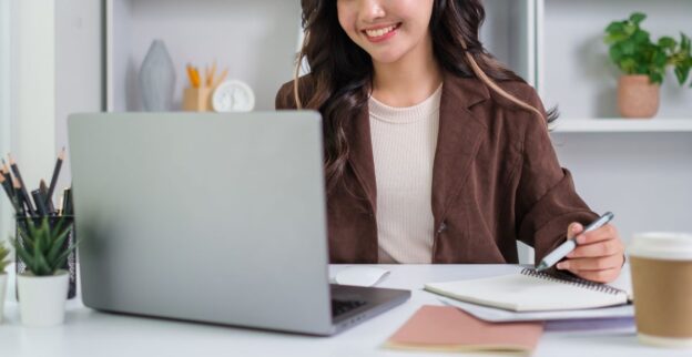 Mulher sorridente trabalhando em frente a um laptop, segurando uma caneta e fazendo anotações, representando liderança feminina no ambiente de trabalho.