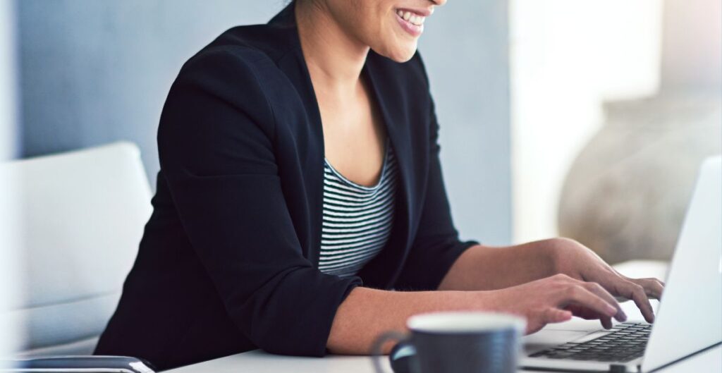 Mulher sorridente usando blazer preto enquanto trabalha em um laptop, representando inovação e empreendedorismo no mercado competitivo.