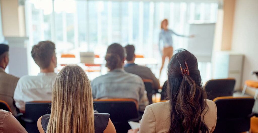 Pessoas em um treinamento empresarial, com uma mulher palestrando ao fundo em uma sala iluminada. Representa a importância de capacitação para equipes e empreendedores.