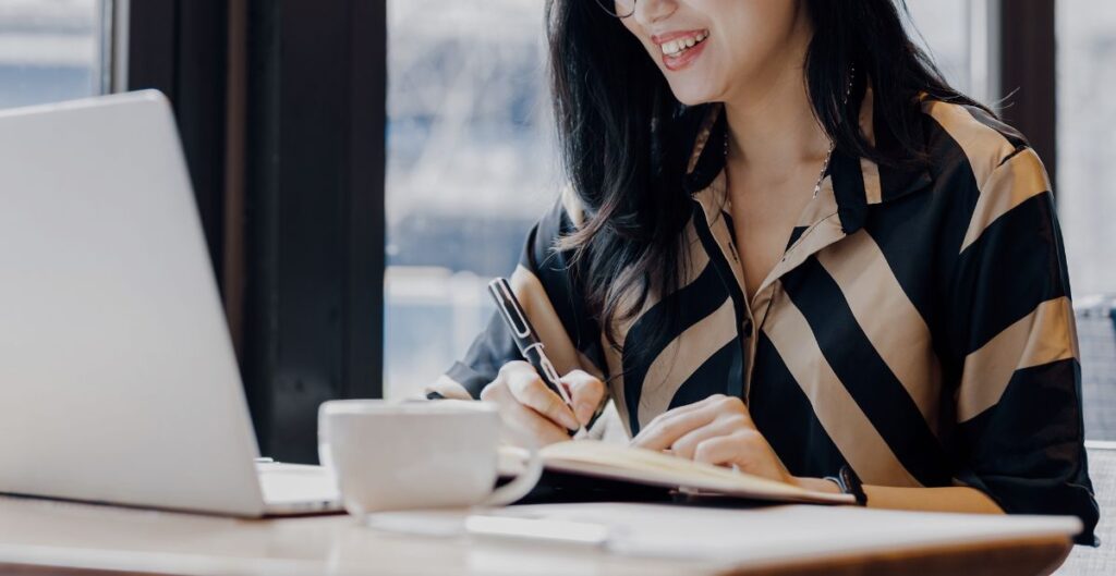 Mulher sorrindo enquanto escreve em um caderno, ao lado de um laptop e uma xícara de café, simbolizando foco e trabalho em liderança feminina.