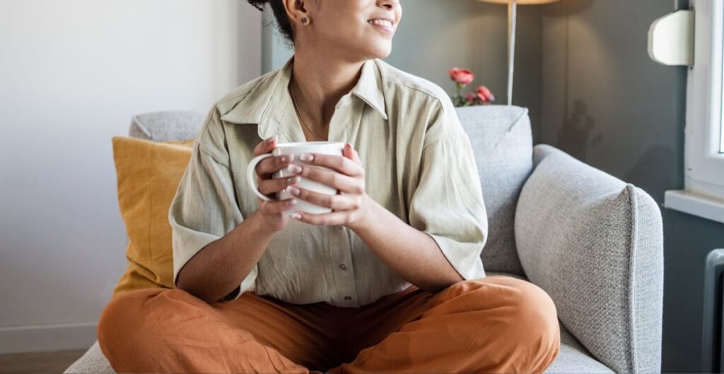 Mulher sentada no sofá, segurando uma xícara de chá ou café, sorrindo e apreciando um momento de tranquilidade.