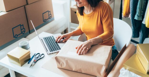 Mulher empreendedora embalando produtos enquanto trabalha em seu laptop, representando o impacto positivo de pequenos negócios