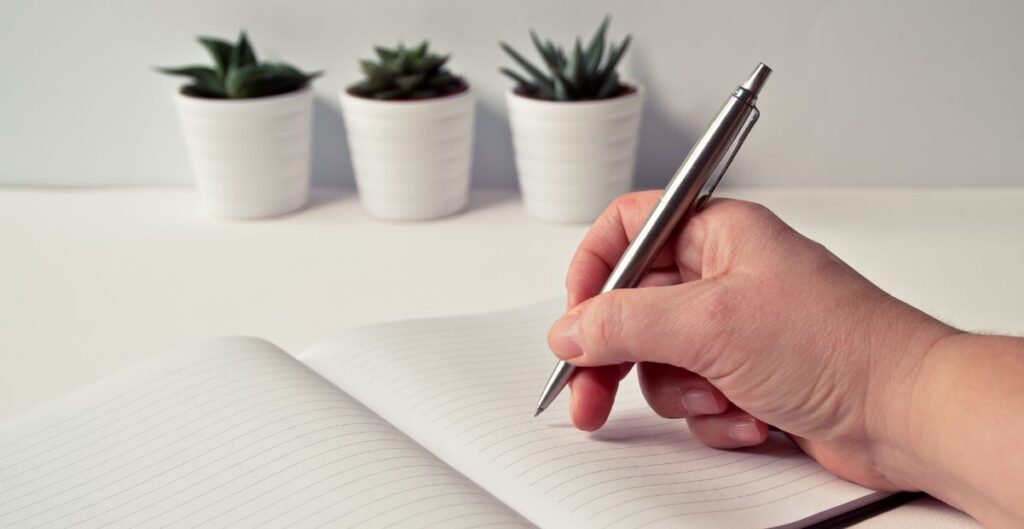 Pessoa escrevendo em caderno sobre mesa branca com vasos de suculentas ao fundo.