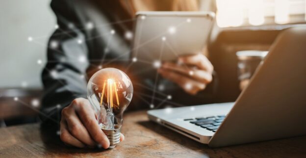 Empresária segurando lâmpada acesa, símbolo de inovação, com notebook e tablet, representando estratégias para empresas se manterem competitivas no mercado digital.