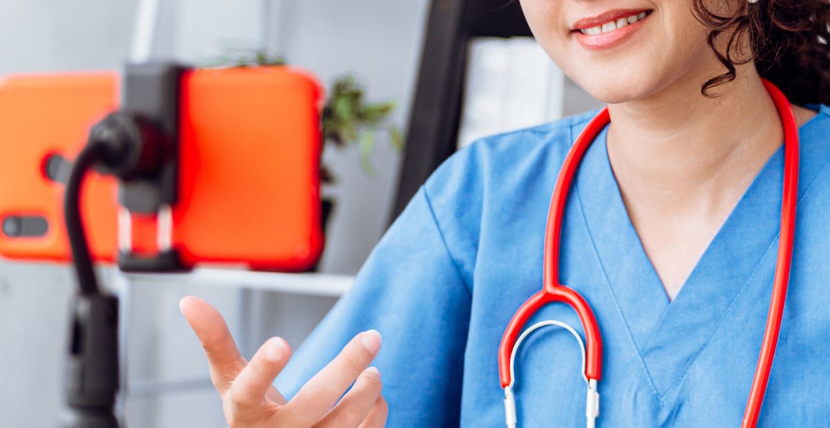 Médica com jaleco azul e estetoscópio grava vídeo para redes sociais com celular em suporte.