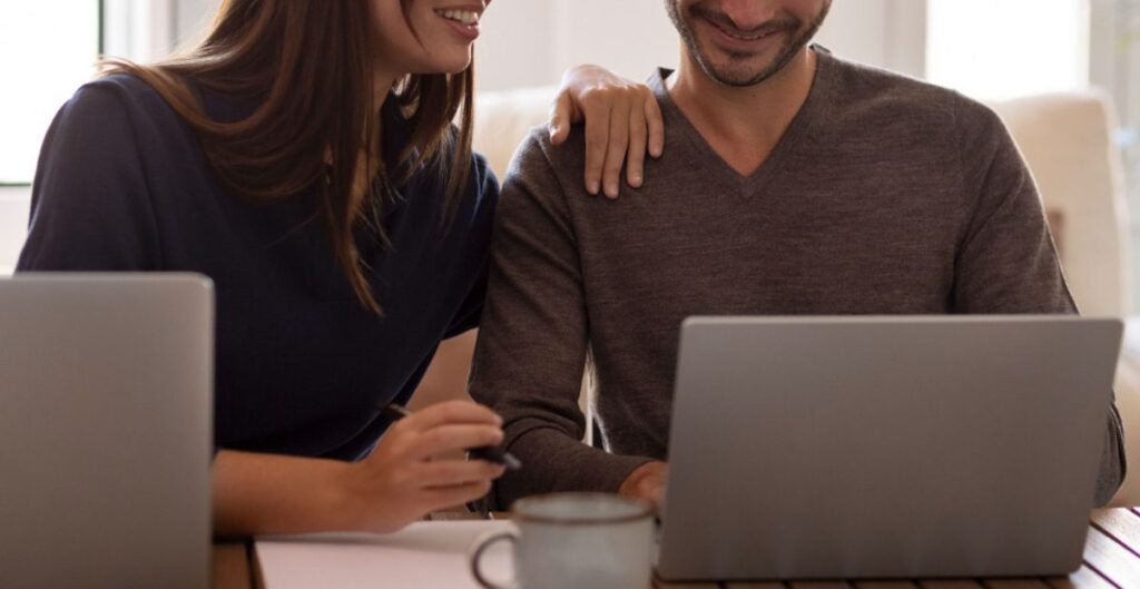 Casal sorridente trabalhando lado a lado em notebooks representando relacionamento amoroso no ambiente de trabalho