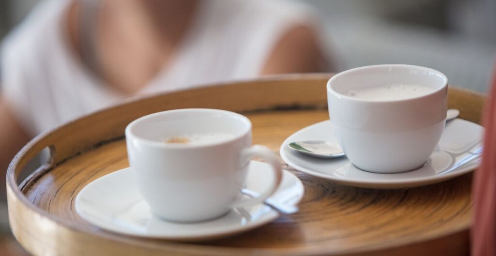 Duas xícaras de café com espuma servidas em pires brancos sobre uma bandeja de madeira, representando diferentes formas de servir café.